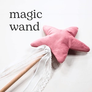 Pode incluir: Uma varinha mágica em forma de estrela rosa com um bastão de madeira e fitas de renda branca. A varinha está sobre uma superfície branca e o texto "magic wand" está acima.