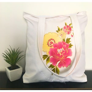 Peut inclure: Sac fourre-tout blanc avec un motif floral. Le sac présente une grande pivoine rose et des fleurs jaunes avec des feuilles vertes. Le sac fourre-tout a deux sangles blanches et est fait d'un tissu blanc doux. Le sac est posé sur une surface noire.