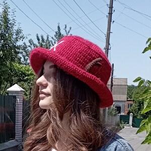May include: A red crocheted hat with a white cross on the front and brown accents. The hat has a wide brim and is likely handmade. The hat is a cosplay item or a character-inspired accessory.