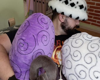 May include: A person wearing a white and black patterned hat is holding a purple and a white plush toy. The toys have a swirl pattern.