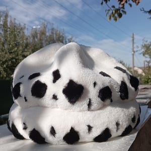 Peut inclure: Un chapeau en peluche blanc et noir à imprimé vache. Le chapeau est fait d'un matériau doux et duveteux et a une forme ronde.