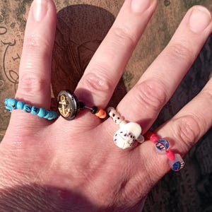 May include: A hand displaying four unique rings. One ring features turquoise beads, another has a black button with a gold anchor, and the others have decorative beads in various colors and designs.