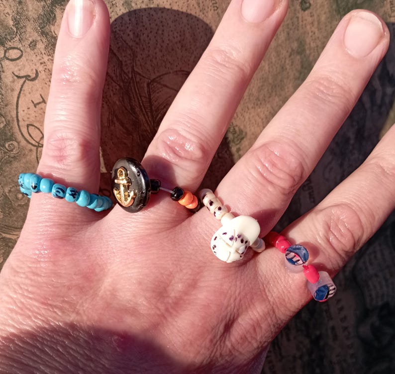 May include: A hand displaying four unique rings. One ring features turquoise beads, another has a black button with a gold anchor, and the others have decorative beads in various colors and designs.