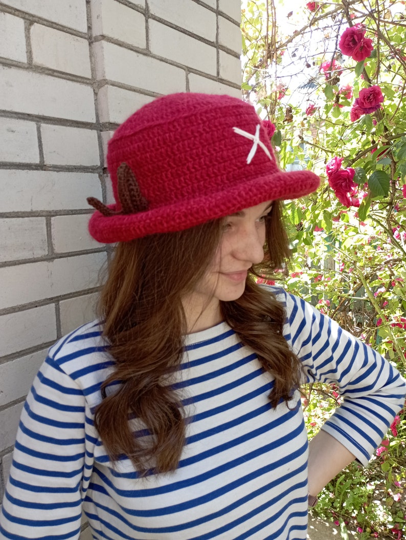 May include: A handmade, red crochet hat with a white cross and brown ear-like accents. The hat has a wide brim and a rounded crown. The hat is a unique accessory.