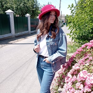 May include: A red crocheted bucket hat with a white cross design. The hat is worn with a denim jacket, white t-shirt, and blue jeans. The background includes a street, green trees, and pink flowers.