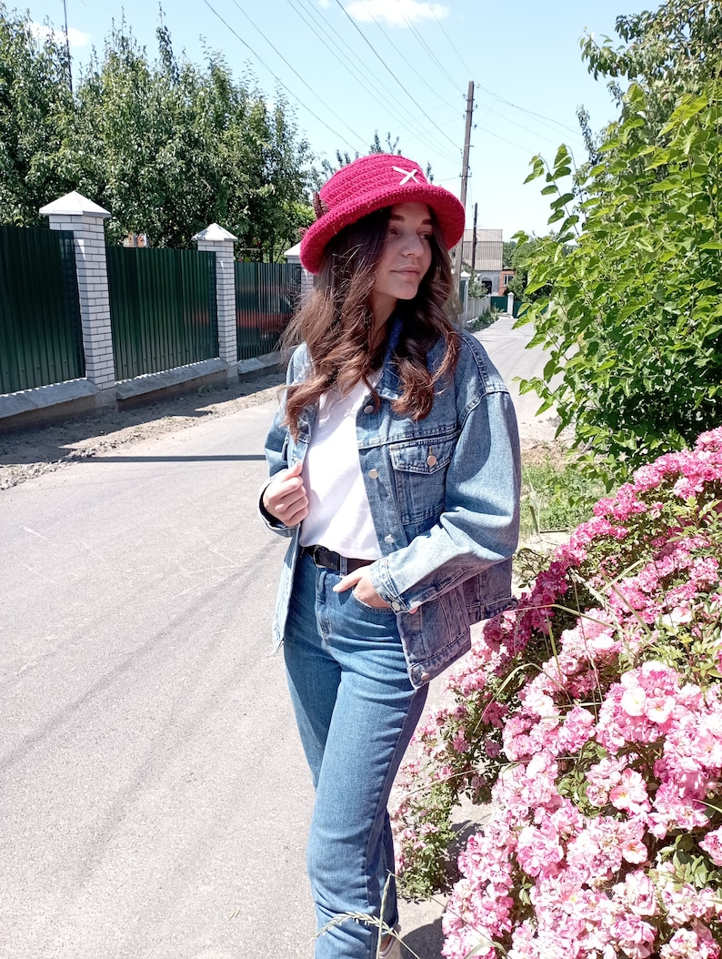 May include: A red crocheted bucket hat with a white cross design. The hat is worn with a denim jacket, white t-shirt, and blue jeans. The background includes a street, green trees, and pink flowers.
