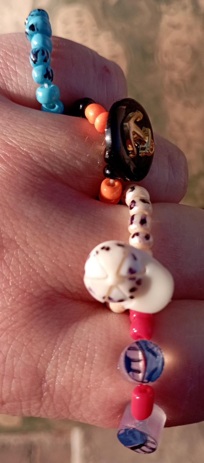 May include: A beaded bracelet with a nautical theme. The bracelet features blue, orange, black, and white beads. A black button with a gold anchor design is a focal point. The bracelet is approximately 7 inches in length.