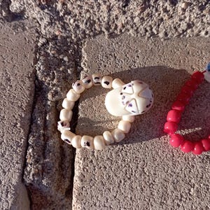 May include: A close-up of a handmade bracelet with white beads and a decorative skull-shaped charm. The skull has purple accents. A second bracelet with red beads is also visible.