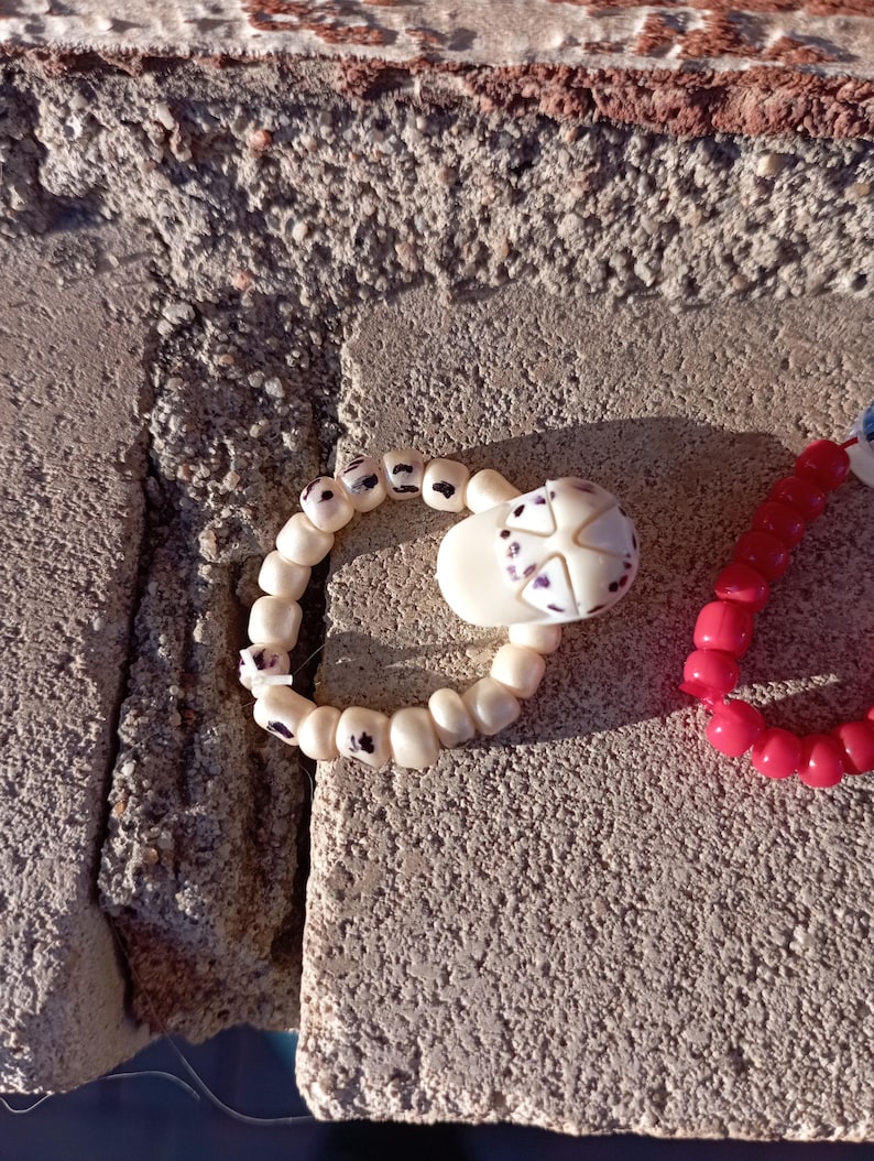 May include: A close-up of a handmade bracelet with white beads and a decorative skull-shaped charm. The skull has purple accents. A second bracelet with red beads is also visible.
