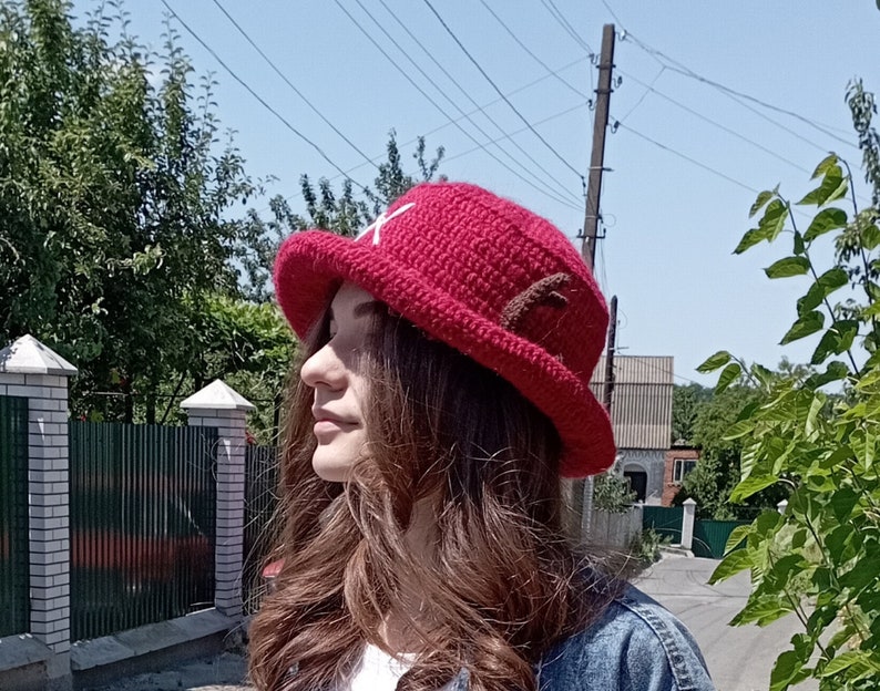 May include: A red crocheted hat with a white cross on the front and brown accents. The hat has a wide brim and is likely handmade. The hat is a cosplay item or a character-inspired accessory.