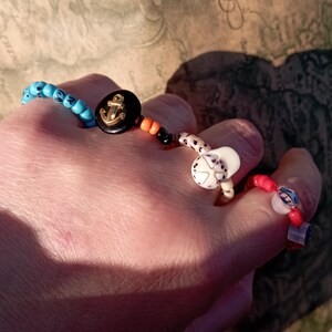 May include: A collection of beaded rings and bracelets. One bracelet features turquoise beads and a black button with a gold anchor. The rings include white, red, and blue beads with various designs. The jewelry is handmade and colorful.