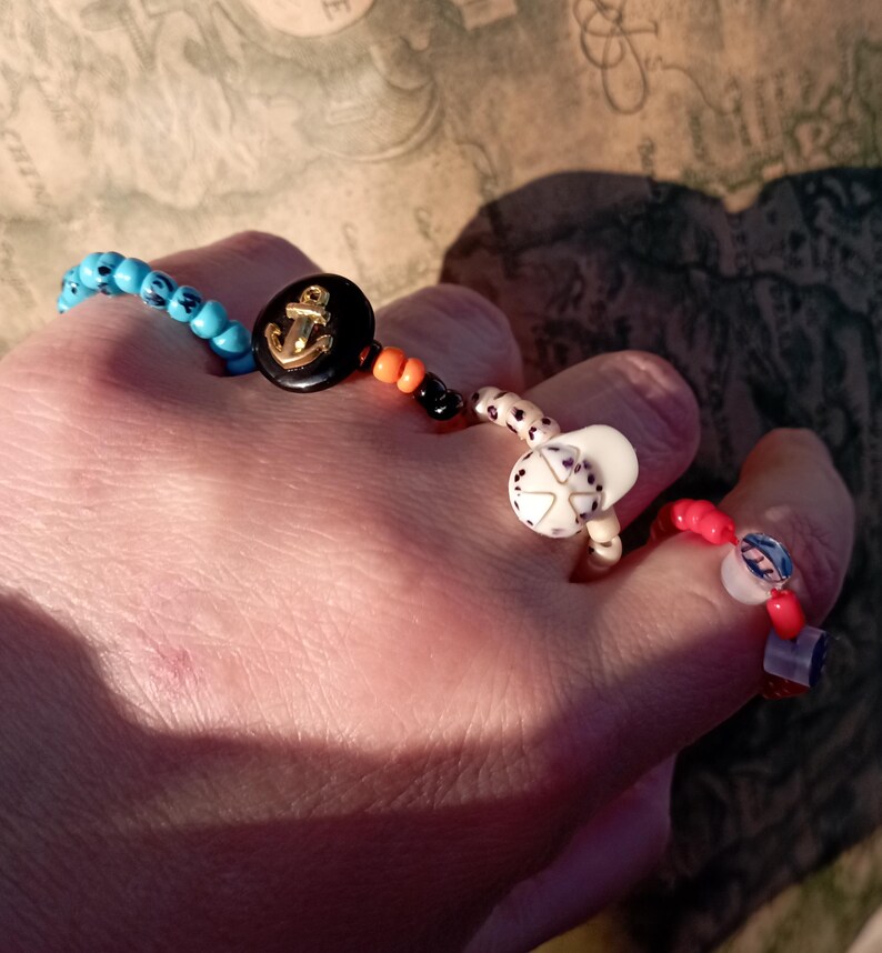 May include: A collection of beaded rings and bracelets. One bracelet features turquoise beads and a black button with a gold anchor. The rings include white, red, and blue beads with various designs. The jewelry is handmade and colorful.
