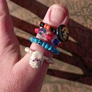 May include: A collection of handmade rings in various styles and colors. One ring features a white skull design, while others incorporate beads in black, orange, red, blue, and clear. A black ring with a gold anchor design is also visible.
