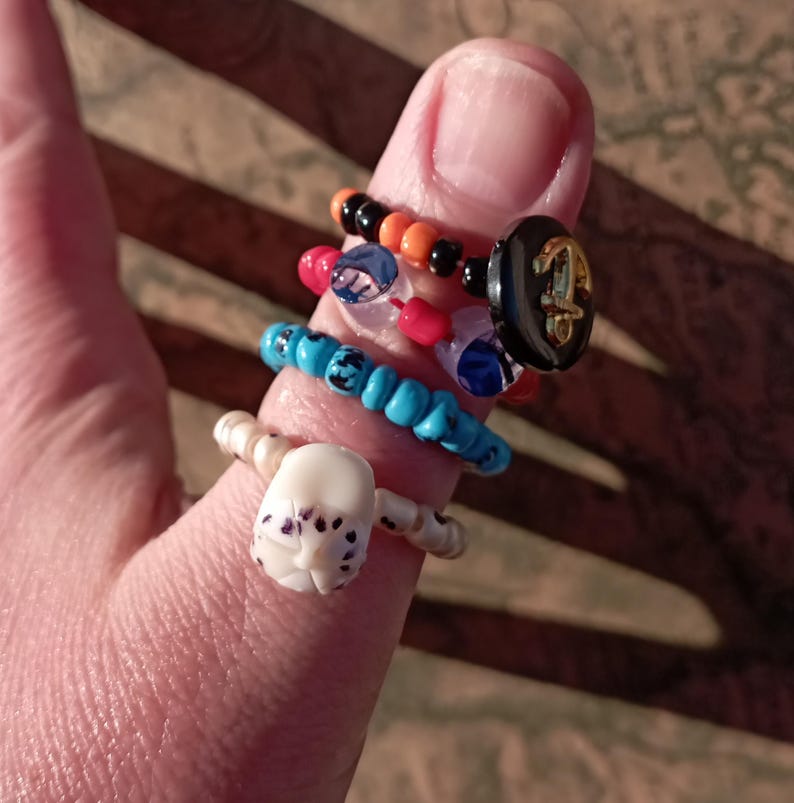 May include: A collection of handmade rings in various styles and colors. One ring features a white skull design, while others incorporate beads in black, orange, red, blue, and clear. A black ring with a gold anchor design is also visible.