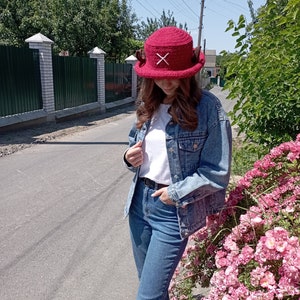 May include: A burgundy crochet hat with a white 'X' design on the front. The hat has a wide brim and two small, brown, horn-like embellishments on the sides. The hat is worn outdoors on a sunny day.