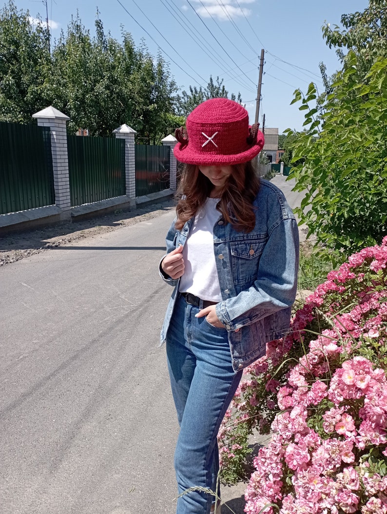 May include: A burgundy crochet hat with a white 'X' design on the front. The hat has a wide brim and two small, brown, horn-like embellishments on the sides. The hat is worn outdoors on a sunny day.