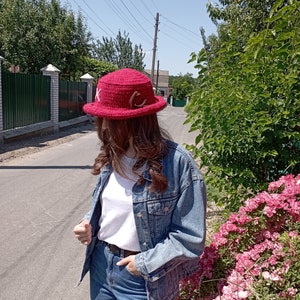 May include: A red crochet hat with a wide brim, adorned with white and brown decorative elements. The hat is a vibrant shade of red, perfect for sun protection. The hat is a stylish accessory.
