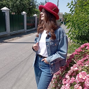 May include: A person wearing a red crochet hat with a white shirt, denim jacket, and blue jeans. The person is standing on a road next to a bush with pink flowers. The background includes a fence and trees.