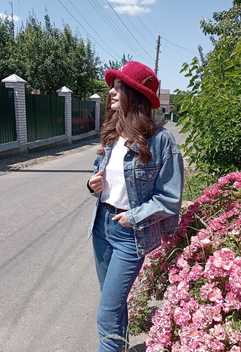 May include: A person wearing a red crochet hat with a white shirt, denim jacket, and blue jeans. The person is standing on a road next to a bush with pink flowers. The background includes a fence and trees.