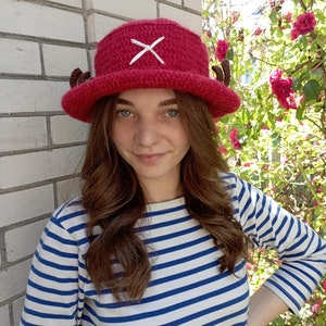 May include: A handmade, red crochet hat with a white cross and small brown horns. The hat is a cosplay item, and is worn with a blue and white striped long-sleeve shirt.