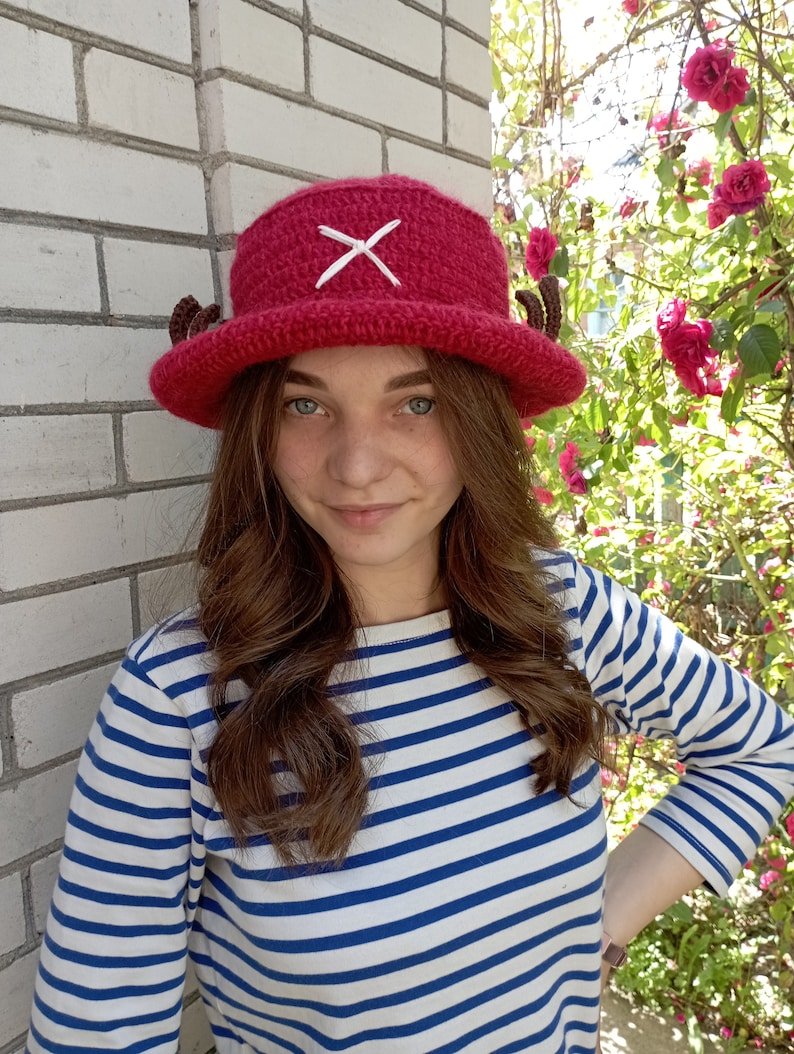 May include: A handmade, red crochet hat with a white cross and small brown horns. The hat is a cosplay item, and is worn with a blue and white striped long-sleeve shirt.