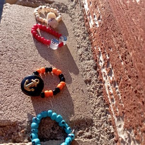 May include: Four beaded rings in a row. One is beige with a white shell, the next is red with clear beads, the third is black and orange with a gold anchor, and the last is blue with black accents. The rings are on a textured surface.