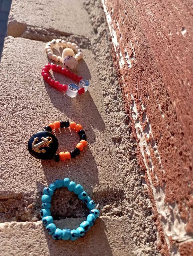 May include: Four beaded rings in a row. One is beige with a white shell, the next is red with clear beads, the third is black and orange with a gold anchor, and the last is blue with black accents. The rings are on a textured surface.