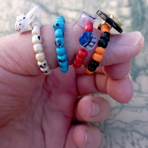 May include: Four beaded rings in various designs. One ring features a white and black spotted dog head. Another has turquoise beads. The third ring has red, blue, and clear beads. The last ring has black, orange, and gold beads.