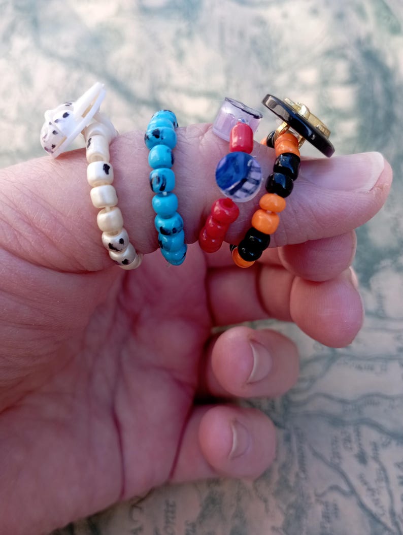 May include: Four beaded rings in various designs. One ring features a white and black spotted dog head. Another has turquoise beads. The third ring has red, blue, and clear beads. The last ring has black, orange, and gold beads.
