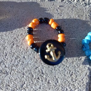 May include: A beaded ring with orange and black beads. A black button with a gold anchor design is attached to the ring. The ring is on a gray, textured surface.