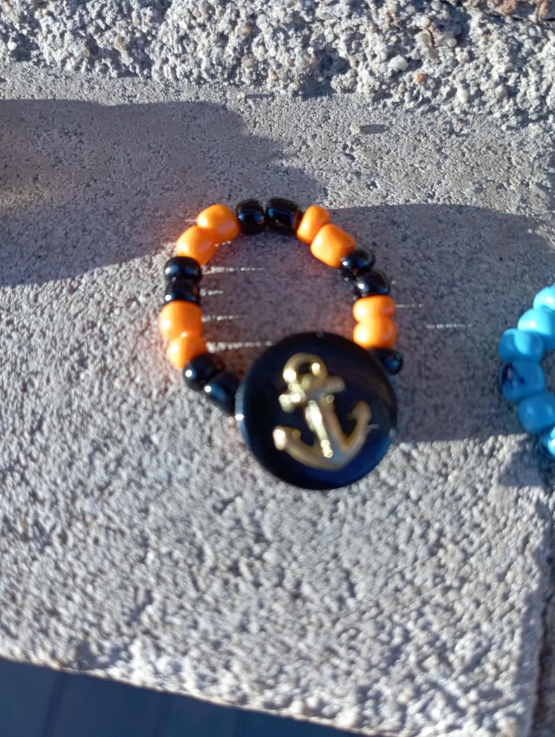 May include: A beaded ring with orange and black beads. A black button with a gold anchor design is attached to the ring. The ring is on a gray, textured surface.