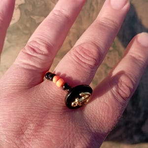 May include: A black and gold ring with an anchor design. The ring features a black base with a gold anchor emblem and is accented with orange and black beads. The ring is on a finger.