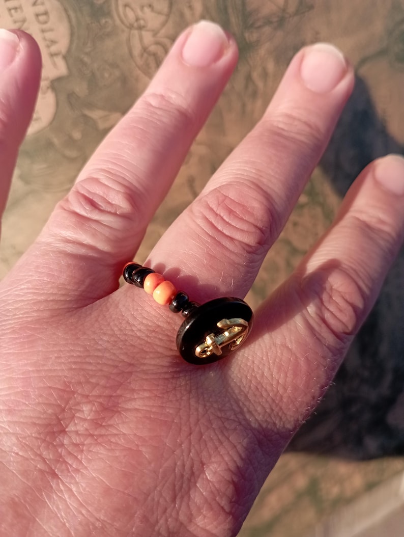 May include: A black and gold ring with an anchor design. The ring features a black base with a gold anchor emblem and is accented with orange and black beads. The ring is on a finger.