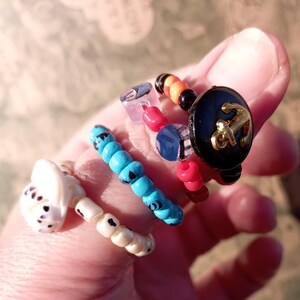 May include: A collection of handmade beaded rings in various designs. One ring features a black button with a gold anchor, another has blue beads, and a third has a white seashell design. The rings are colorful and unique.
