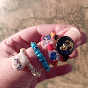 Könnte beinhalten: Eine Auswahl an Ringen mit Perlen in verschiedenen Designs. Ein Ring hat ein weißes, geformtes Design mit schwarzen Punkten. Ein weiterer hat türkisfarbene Perlen, und ein dritter hat rote und klare Perlen. Der letzte Ring hat einen marineblauen Knopf mit einem goldenen Anker.