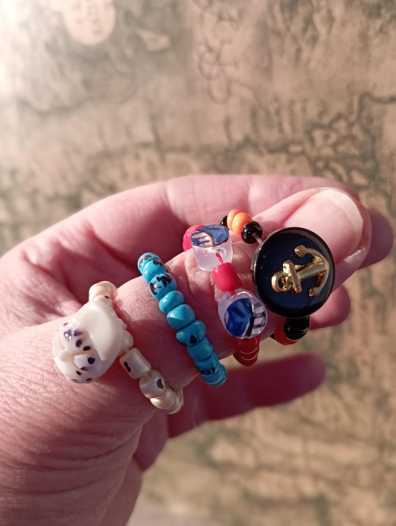 May include: A collection of beaded rings in various designs. One ring features a white, sculpted design with black spots. Another has turquoise beads, and a third has red and clear beads. The last ring has a navy blue button with a gold anchor.
