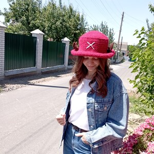May include: A burgundy crochet hat with a wide brim, featuring a white 'X' design and small, brown decorative elements. The hat is a unique accessory, perfect for adding a touch of handmade charm to any outfit.