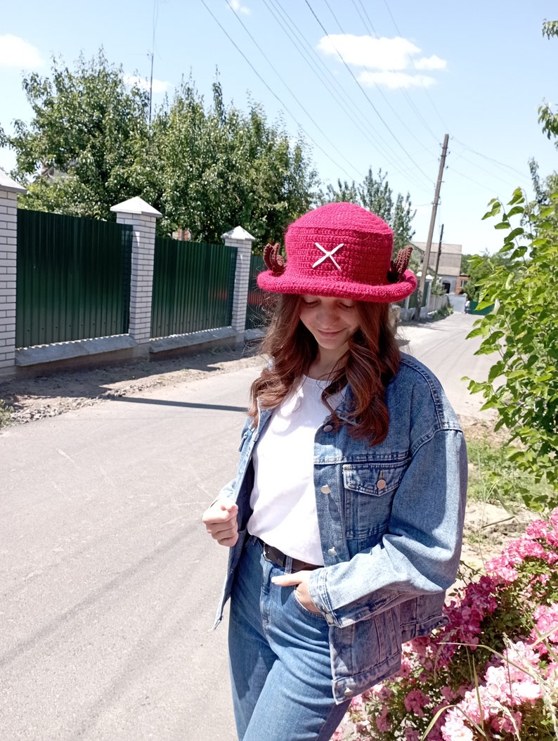 May include: A burgundy crochet hat with a wide brim, featuring a white 'X' design and small, brown decorative elements. The hat is a unique accessory, perfect for adding a touch of handmade charm to any outfit.