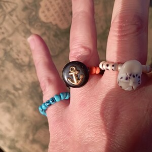 May include: A hand wearing four beaded rings. One ring is turquoise, another has a black disc with a gold anchor, and a third has a white, spotted bead. The fourth ring has orange and white beads.