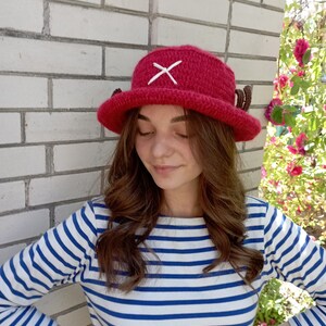 May include: A handmade, red crochet hat with a white cross on the front and small brown antlers on the sides. The hat is a bucket style, perfect for cosplay or everyday wear. The hat is shown in front of a white brick wall.