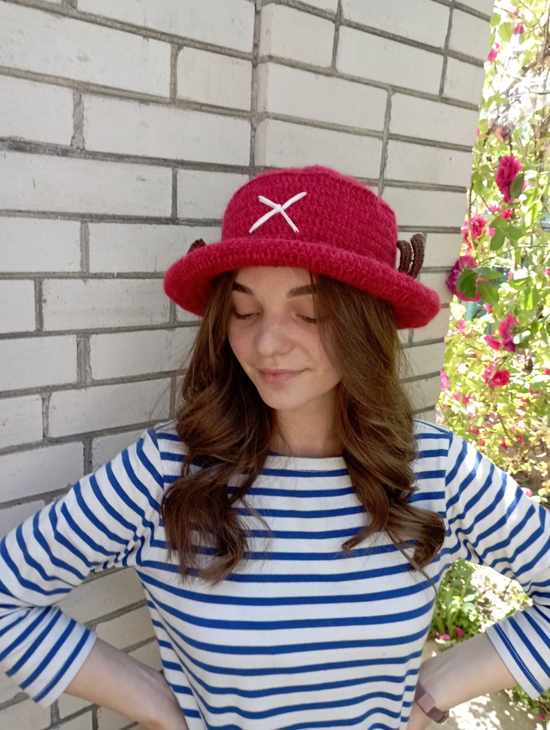May include: A handmade, red crochet hat with a white cross on the front and small brown antlers on the sides. The hat is a bucket style, perfect for cosplay or everyday wear. The hat is shown in front of a white brick wall.