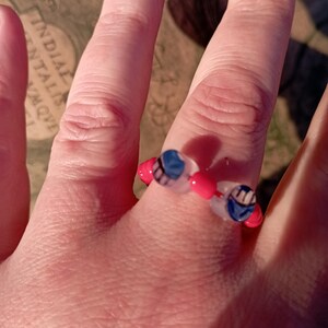 May include: A beaded ring featuring a design with red, white, and blue beads. The ring is composed of small, round beads strung together to form a circular shape. The ring is worn on a finger.