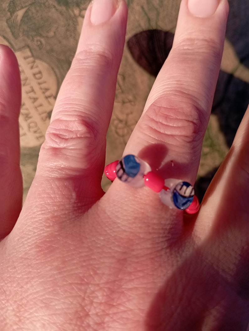 May include: A beaded ring featuring a design with red, white, and blue beads. The ring is composed of small, round beads strung together to form a circular shape. The ring is worn on a finger.