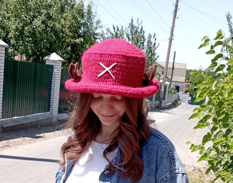 May include: A handmade, magenta crochet hat with a white cross design on the front. The hat features two brown, antler-like embellishments on the sides. This unique hat is a fun accessory for cosplay or everyday wear.