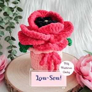 May include: A handmade, plush, red poppy flower with a black center and green leaves, displayed in a pink pot. The text "Low-Sew!" is visible on a pink rectangle. The item is placed on a wooden surface.