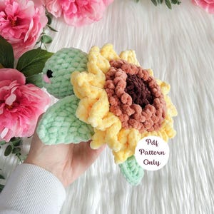 May include: A crochet turtle plush with a light green head and flippers, featuring a shell designed as a yellow and brown sunflower. A white circular tag is attached to the shell with the text Pdf Pattern Only printed in dark brown letters.