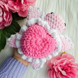 May include: A pink and white crocheted stuffed animal shaped like a turtle. The turtle has a heart-shaped shell and a white ruffle around the edge.