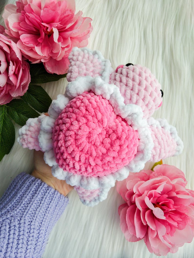 May include: A pink and white crocheted stuffed animal shaped like a turtle. The turtle has a heart-shaped shell and a white ruffle around the edge.