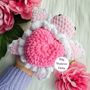 Puede incluir: Juguete amigurumi de crochet rosa y blanco con forma de corazón y alas. El juguete está siendo sostenido en una mano. El texto "Pdf Pattern Only" está en un círculo blanco en la esquina inferior derecha de la imagen.