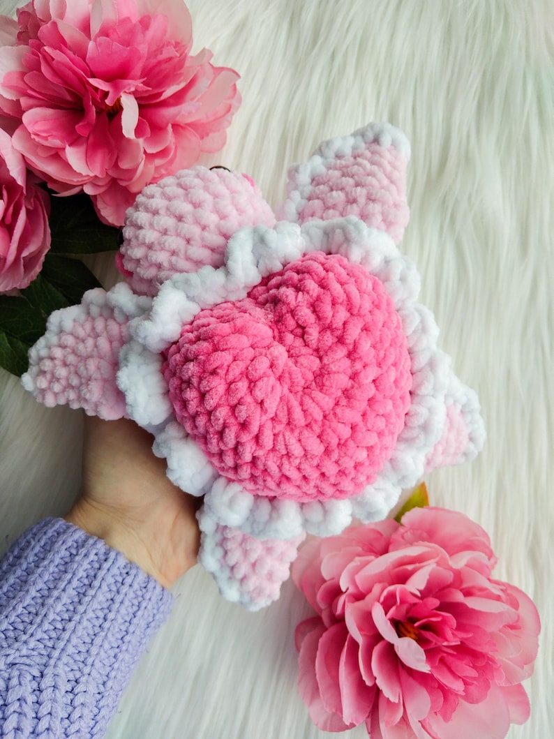 May include: A pink and white crocheted heart-shaped turtle plush toy. The toy is held in a hand with a purple sweater.